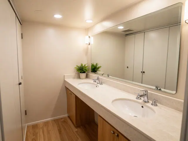 Inside a restroom trailer: a long light stone vanity with two sinks and chrome faucets, a wide mirror, two small potted plants, wood-look flooring and warm recessed lighting.